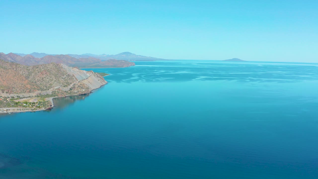 Loreto coastal aerial with calm blue water in the foreground, the hilly shore receding from the left, pale ripples across the bay, distant landforms near the horizon, and no visible people.