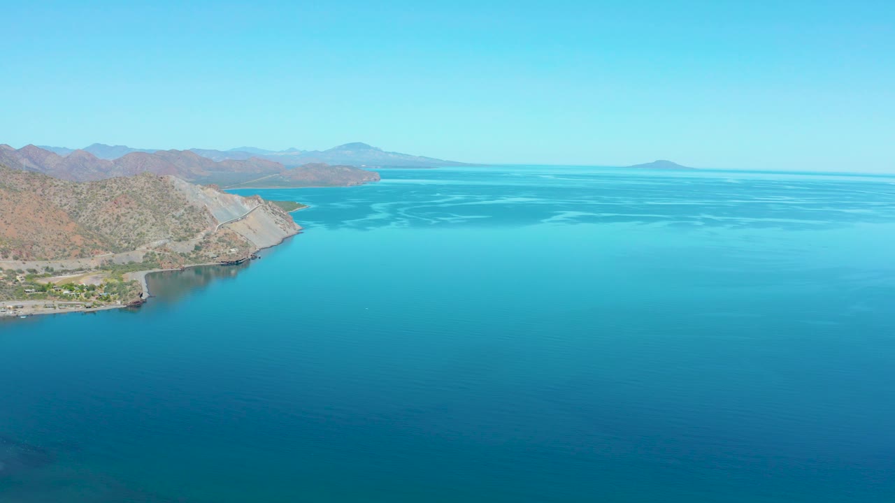 Aerial coastline view with road, small structures, hills, and blue water.