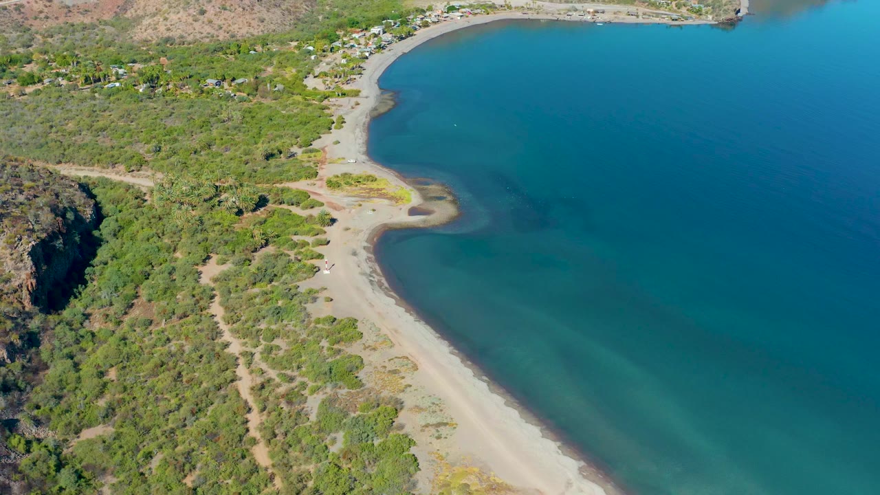 Aerial frame from Loreto, Baja California Sur showing a curved sandy beach, turquoise ocean, green coastal vegetation, a rocky left edge, and no visible people.