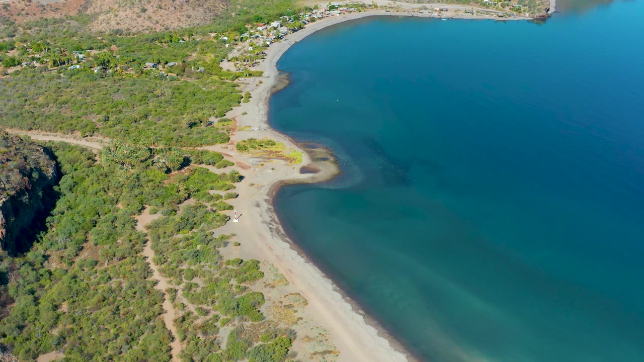 High aerial coastal view with turquoise shallows, sandy shore, scrub vegetation, and road.