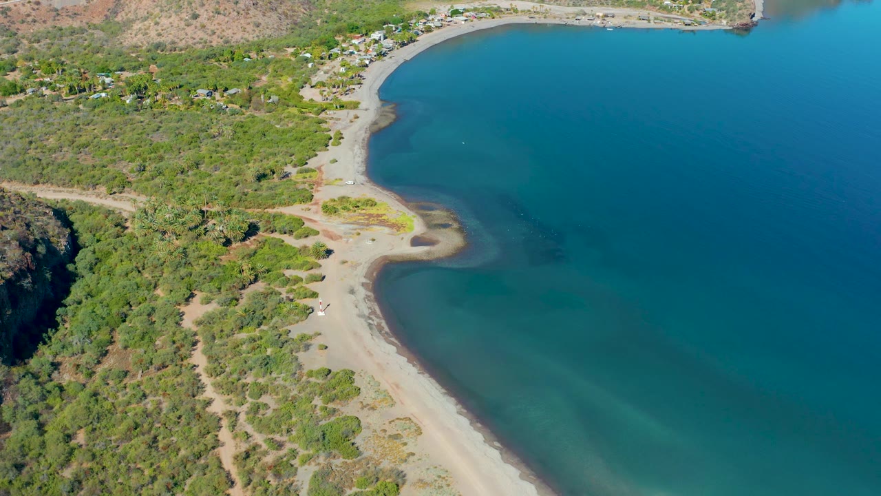 Aerial view of the Loreto bay shoreline with broad blue water to the right, a curved sandy beach, scrub vegetation, small distant structures, and no visible people.