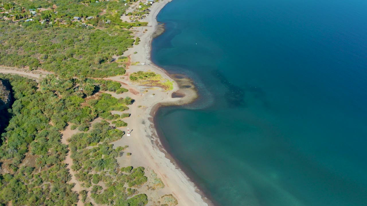 Aerial view over Loreto, Baja California Sur, showing a pale sandy beach beside clear blue water, dense green vegetation inland, and a small vehicle on the beach with no visible people.