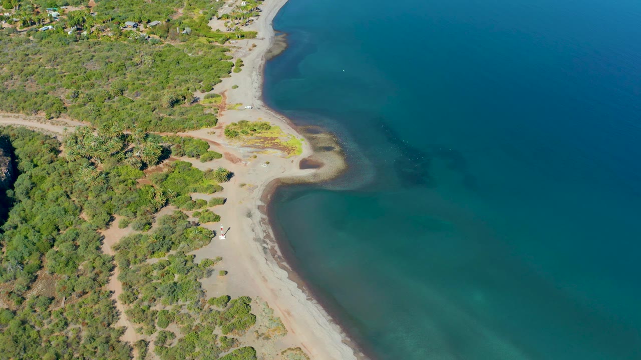 Aerial view in Loreto, Baja California Sur, with more open blue water filling the right side, a pale beach running diagonally, dense greenery at left, and the small vehicle on the lower sand with no visible people.