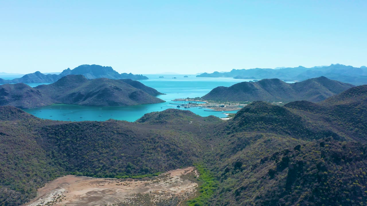 Aerial frame of Juncalito, Baja California Sur with rugged foreground hills, a turquoise inlet, small shoreline development, hazy distant landforms, and no visible people.