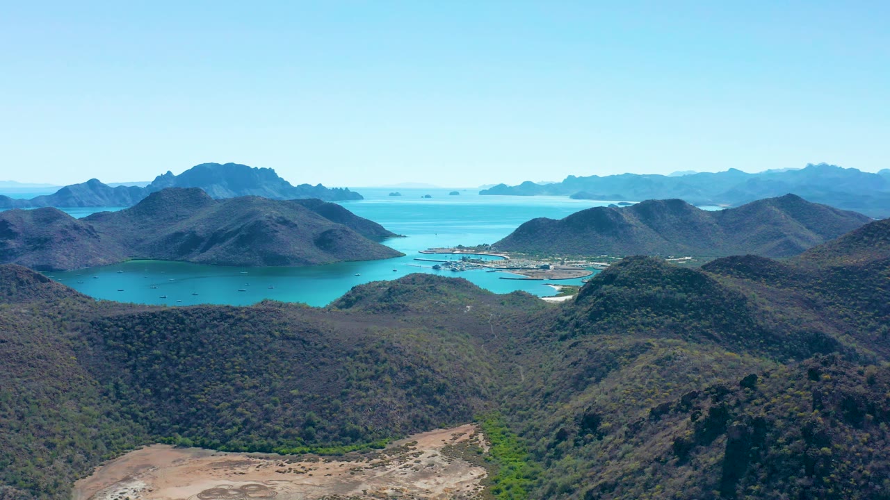Early aerial frame over Juncalito with foreground hillside texture, bright blue bay water, layered mountain ridges, a small shoreline area, and no visible people.