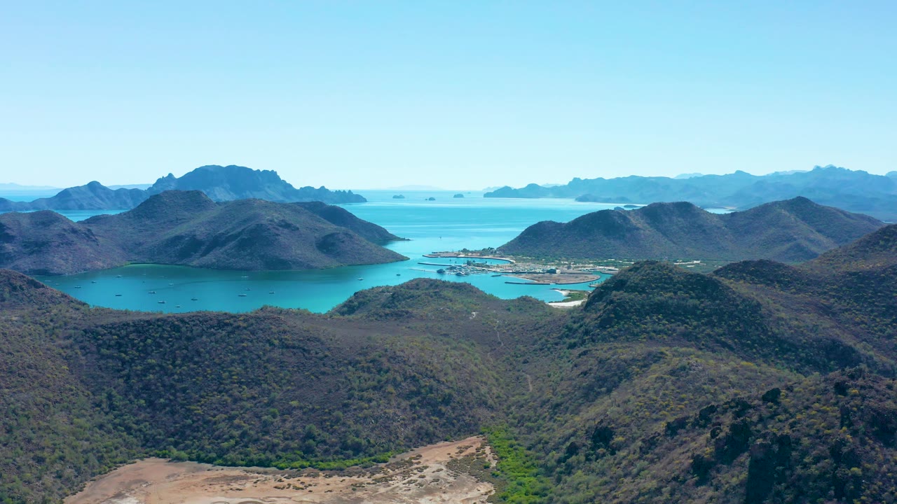 Aerial frame from Juncalito with the central bay partly enclosed by hills, a small developed shoreline strip, distant islands or headlands, clear daylight, and no visible people.