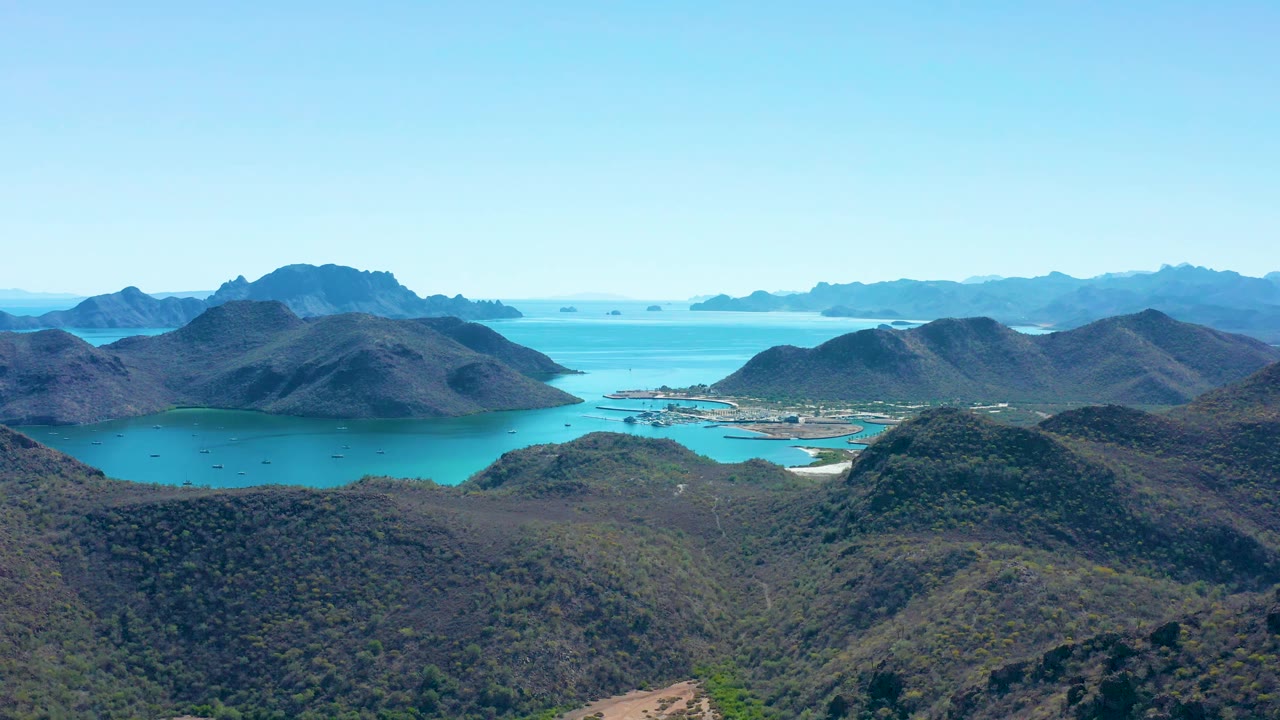 Aerial frame in Juncalito showing the bay held between rounded hills, small dark boat shapes on the water, pale shoreline areas, and no visible people.
