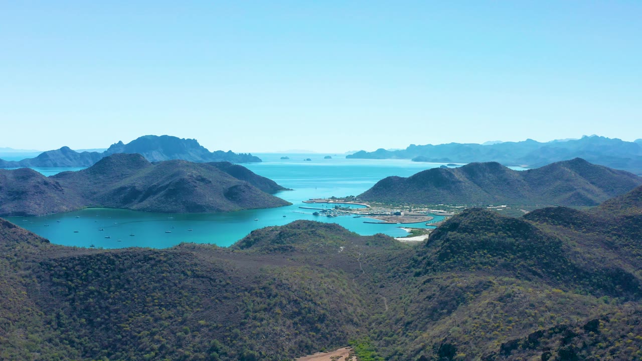High aerial view of Juncalito with textured green-brown hills in the foreground, turquoise water across the middle, island silhouettes on the horizon, and no visible people.