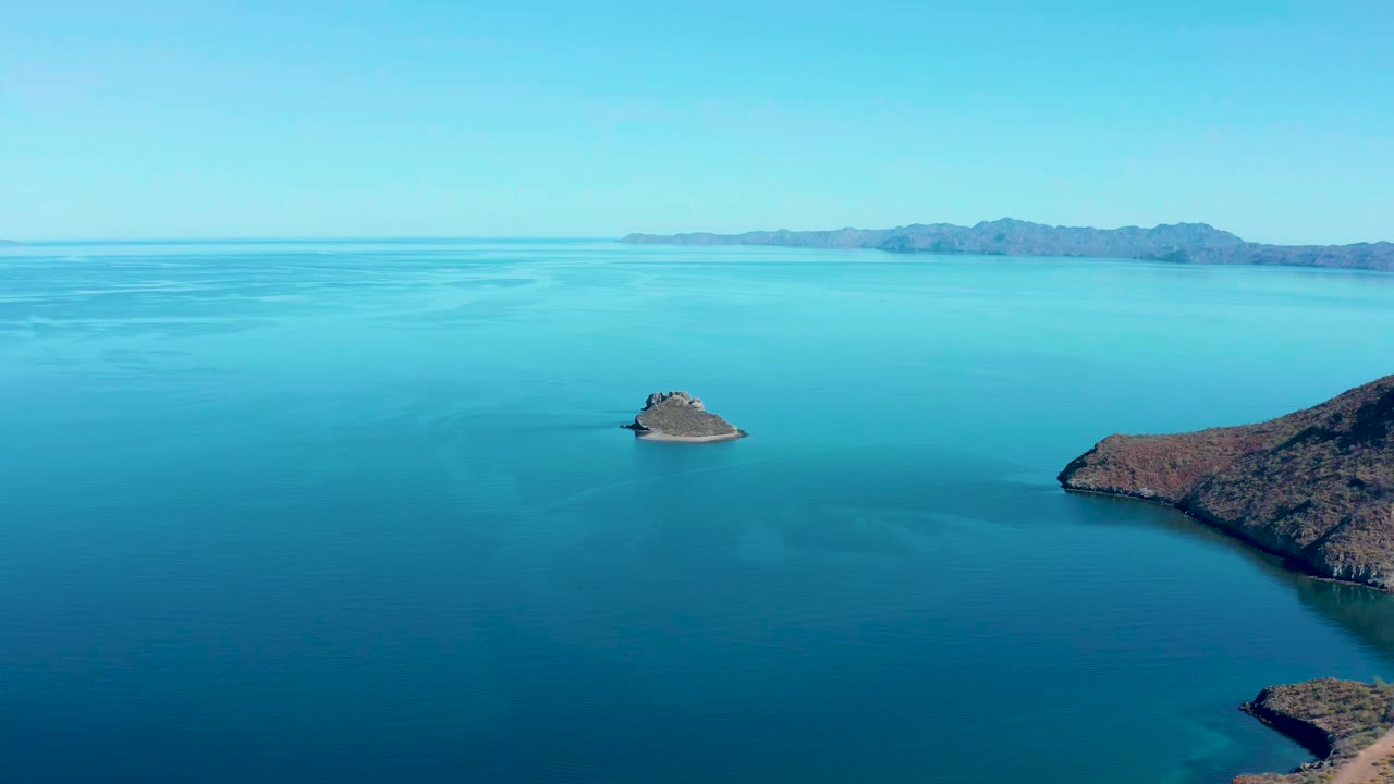 At 0:00 in Juncalito, Baja California Sur, a small rocky island sits in calm blue water with a larger coastal landmass at right, distant hills on the horizon, and no visible people.