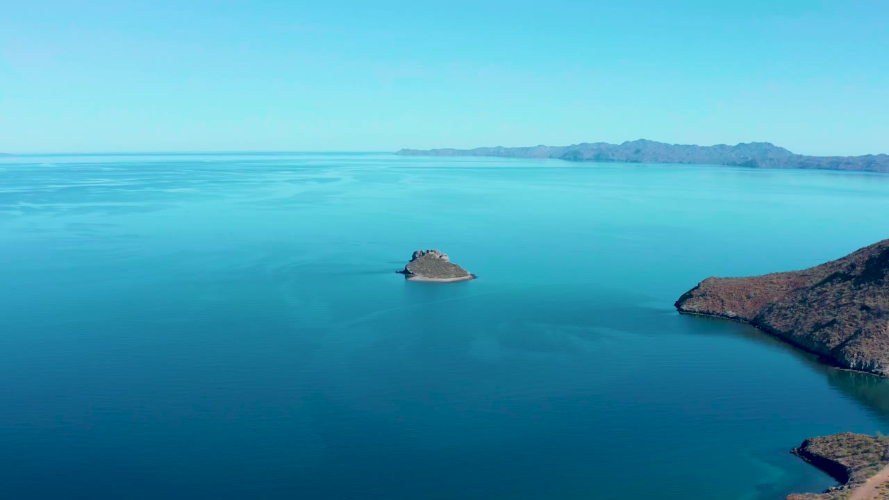 At 0:01, the Juncalito aerial view holds on the rocky islet surrounded by smooth blue ocean, with brown shoreline on the right edge, low hills beyond, and no visible people.
