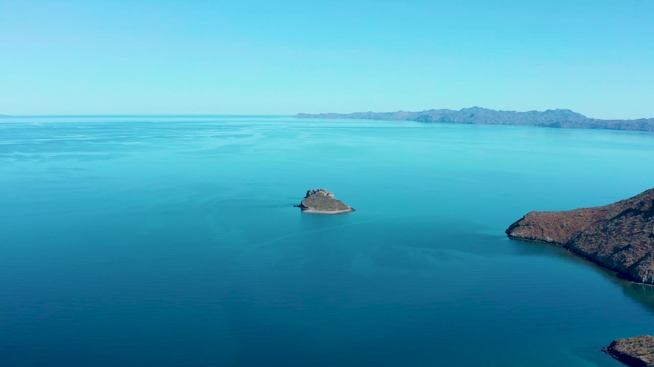 One second into the Juncalito clip, the small rocky island remains in open blue water with the larger right-side landmass closer to the frame edge, distant hills on the horizon, and no visible people.
