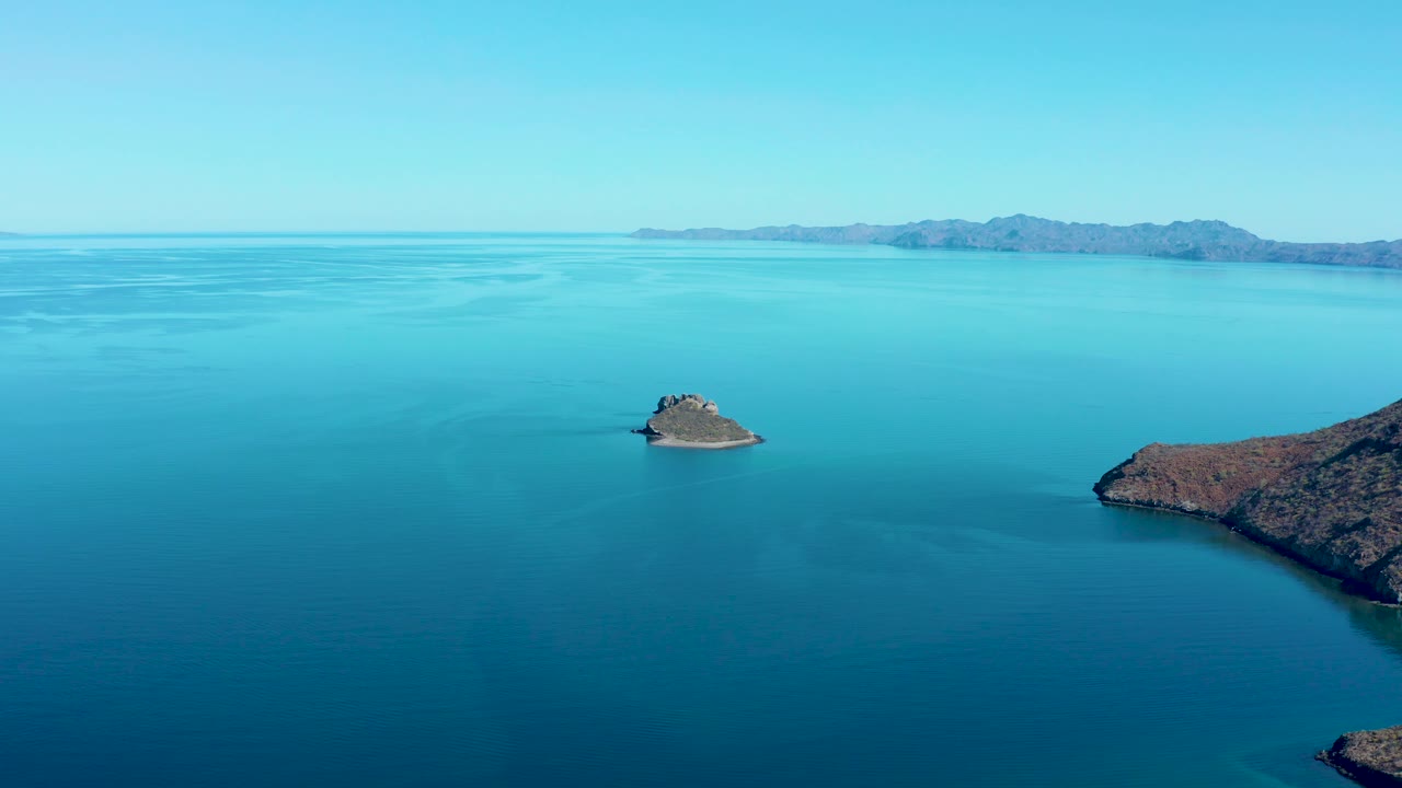 At 0:03, the aerial view keeps the Juncalito island in the midground with broad blue ocean in front, a rocky shoreline on the right, distant hills, a clear sky, and no visible people.