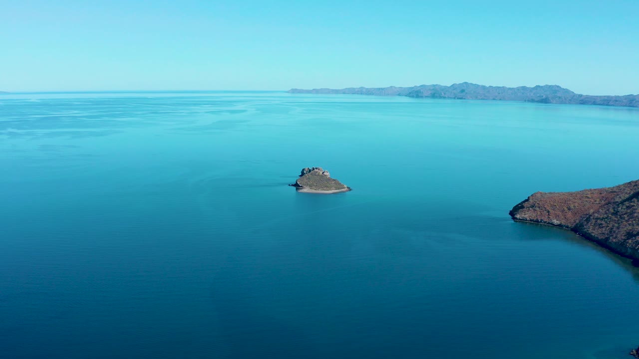 Small rocky island in blue ocean with distant mountains.