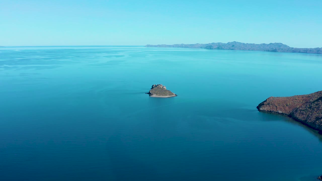 At 0:01, the small island remains centered in blue water, with a reddish coastal point entering from the right and distant mountains across the Juncalito horizon, with no visible people.