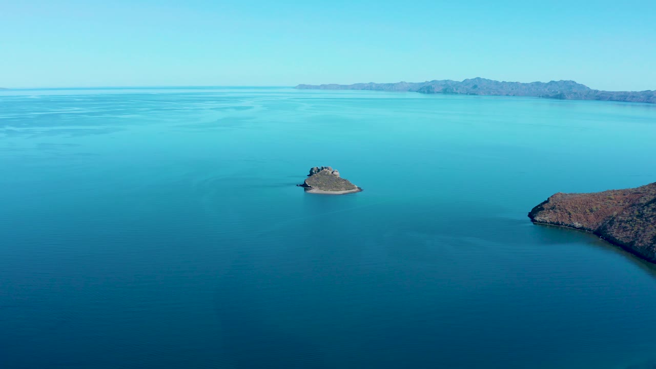 At 0:03, the small rocky island is surrounded by smooth blue water, with the right edge of the coastline and a low mountain range visible in the background, and no visible people.