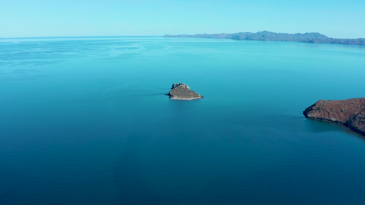 Mid-clip aerial frame of calm Juncalito ocean with the small rocky island below the horizon, faint water ripples, a distant mountain ridge, and no visible people.