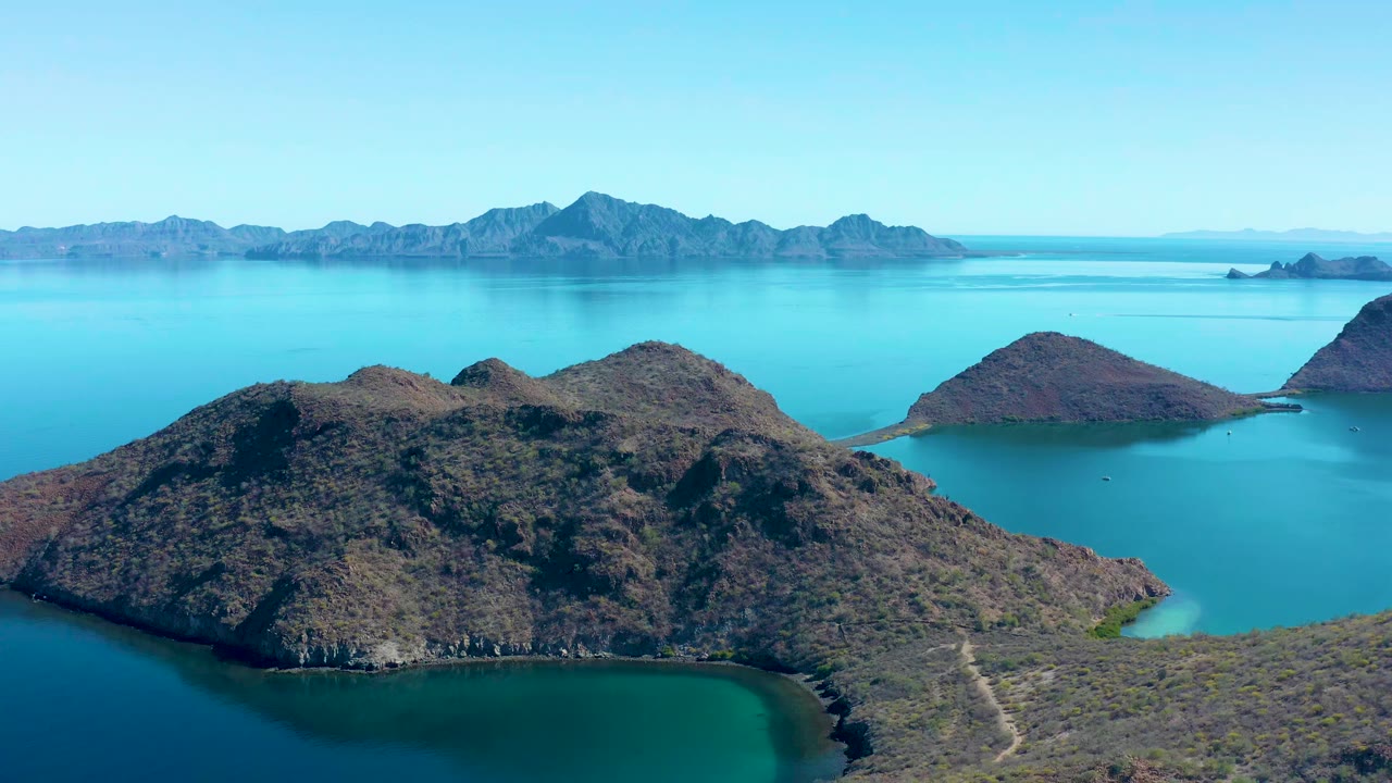 Aerial frame over Juncalito showing scrub-covered rocky slopes in the foreground, a pale turquoise cove to the right, darker blue water below, distant islands, and no visible people.