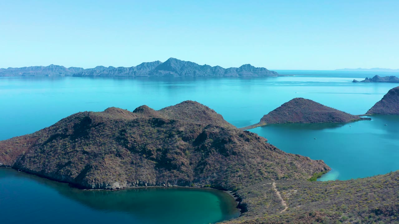Wide aerial frame of Juncalito showing the rocky island ridge slightly more centered, blue-green water in the lower cove, distant mountain silhouettes, clear sky, and no visible people.