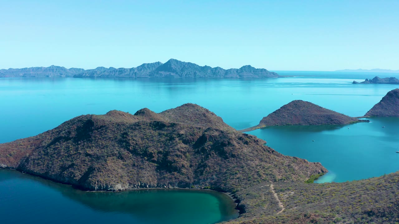 Wide aerial frame over Juncalito with a large rocky island spanning the foreground, a protected turquoise cove below it, small islands beyond, distant mountains, and no visible people.