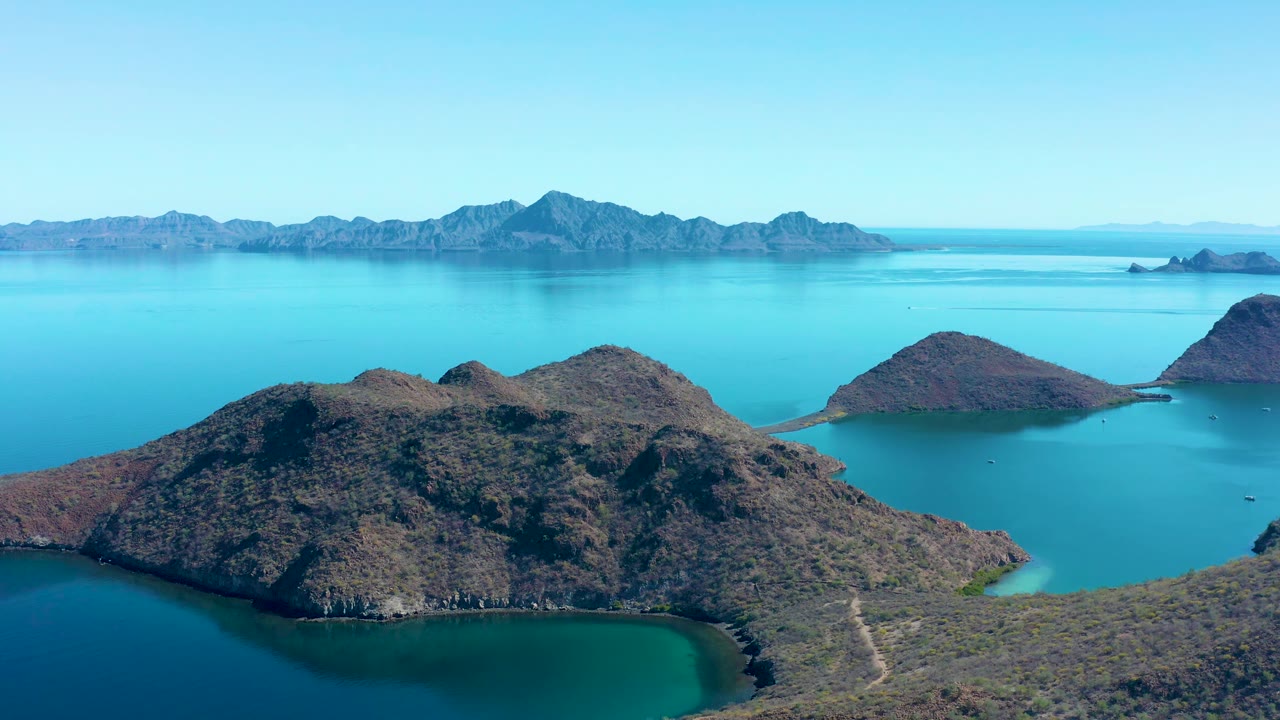 Second aerial frame in Juncalito, Baja California Sur, with the main rocky island centered lower in frame, smaller islands to the right, distant mountains, and no visible people.