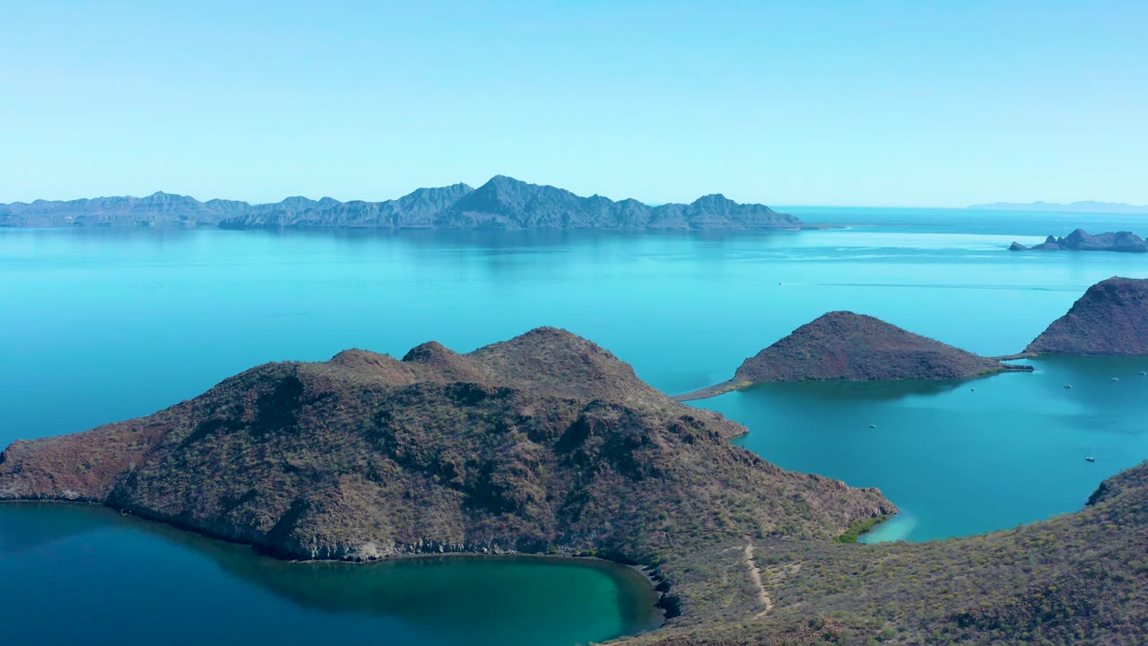 Second-frame aerial composition over Juncalito rocky island slopes, calm blue-green water, a long mountain horizon, clear sky, and no visible people.