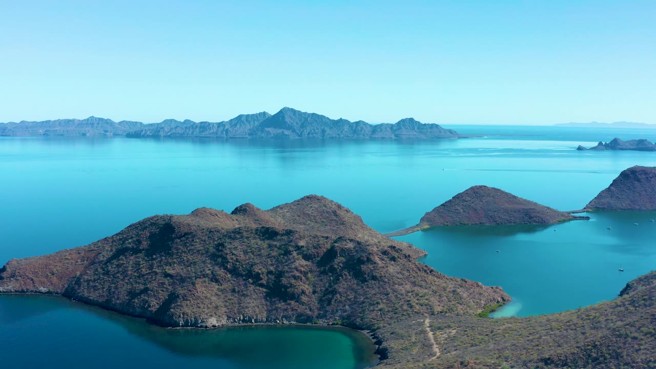 Aerial frame in Juncalito showing dry rocky island terrain in the foreground, clear turquoise water around coves, hazy background mountains, and no visible people.