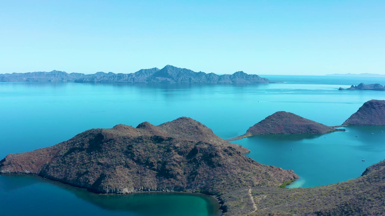 Mid-clip aerial view of Juncalito islands with a rocky foreground ridge, bright turquoise sea, distant ridgelines, clear sky, and no visible people.