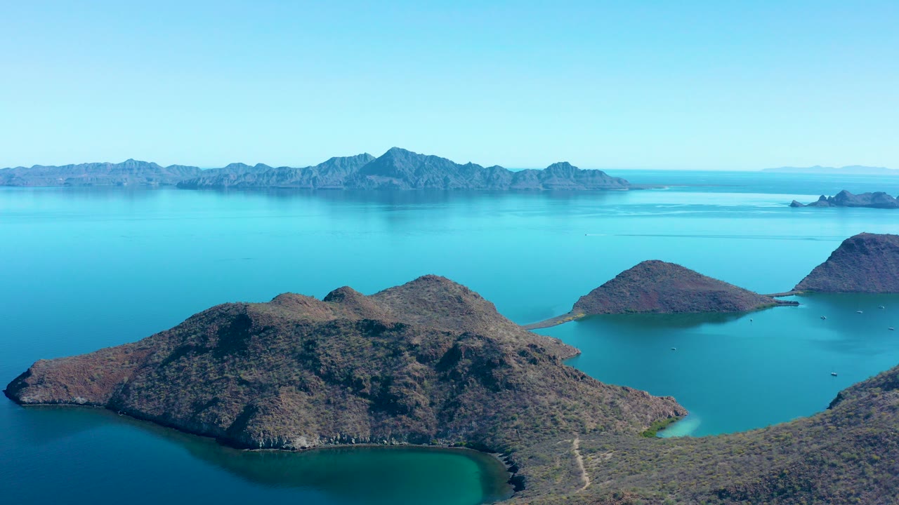 Wide aerial view over rocky islands in Juncalito, Baja California Sur, with turquoise shallows, deeper blue water, distant mountain ridges, and no visible people.