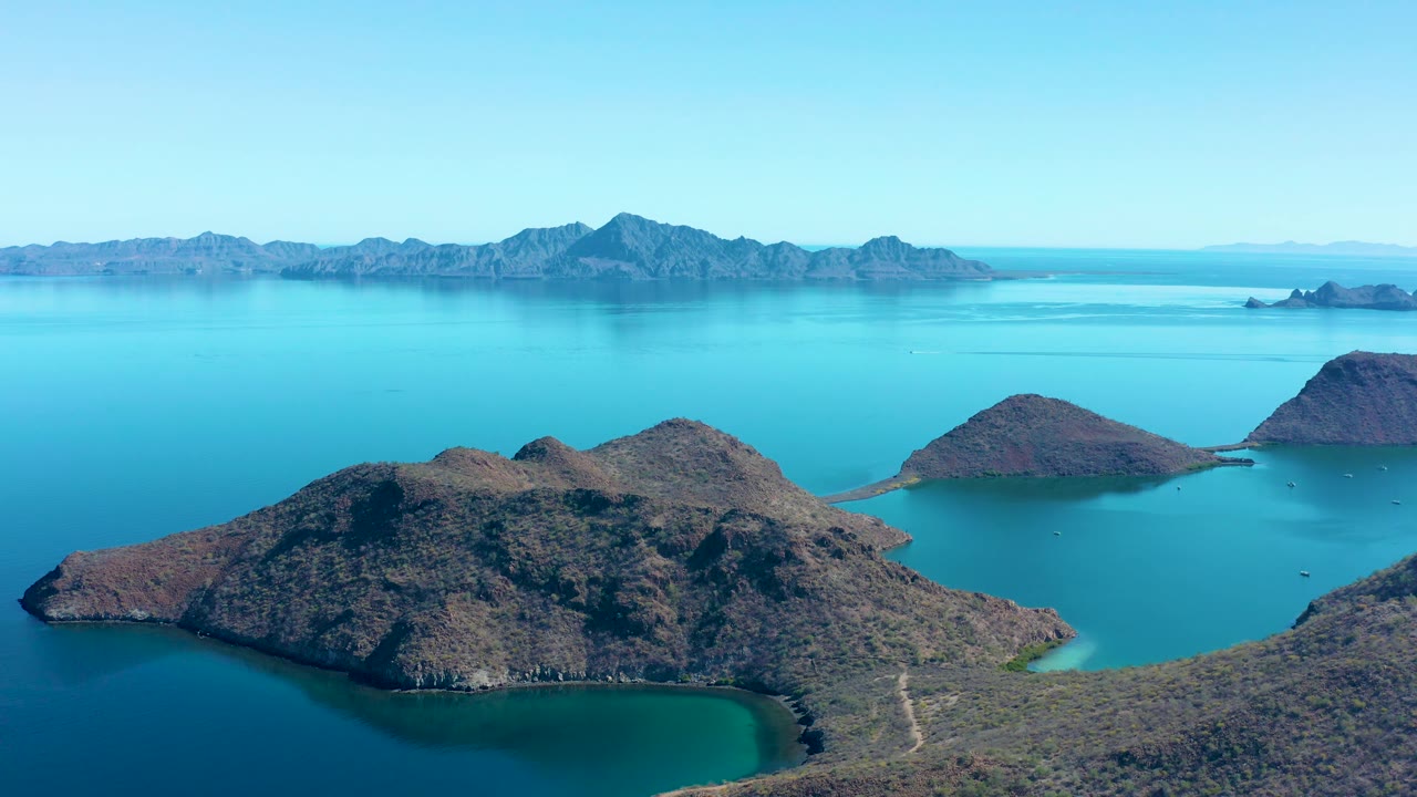 Juncalito aerial frame with rocky islands across calm blue water, pale turquoise edges near shore, distant landmasses, a clear sky, and no visible people.
