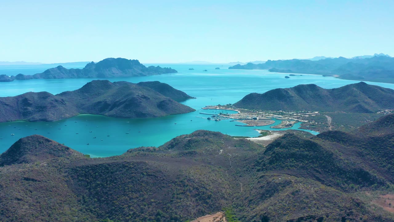 Wide aerial frame of Juncalito showing green-brown hills around clear blue bay water, a compact coastal settlement and harbor, layered islands on the horizon, and no visible people.