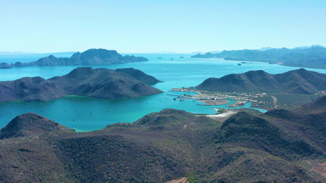 Wide drone view of mountain-ringed bay with boats and shoreline buildings.