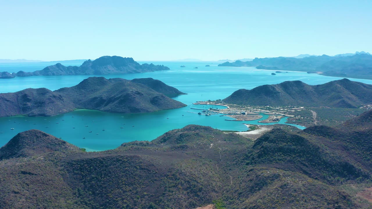 Aerial frame over Juncalito with green-brown hills in the foreground, pale roads and shoreline buildings in the midground, boats in the bay, and no visible people.