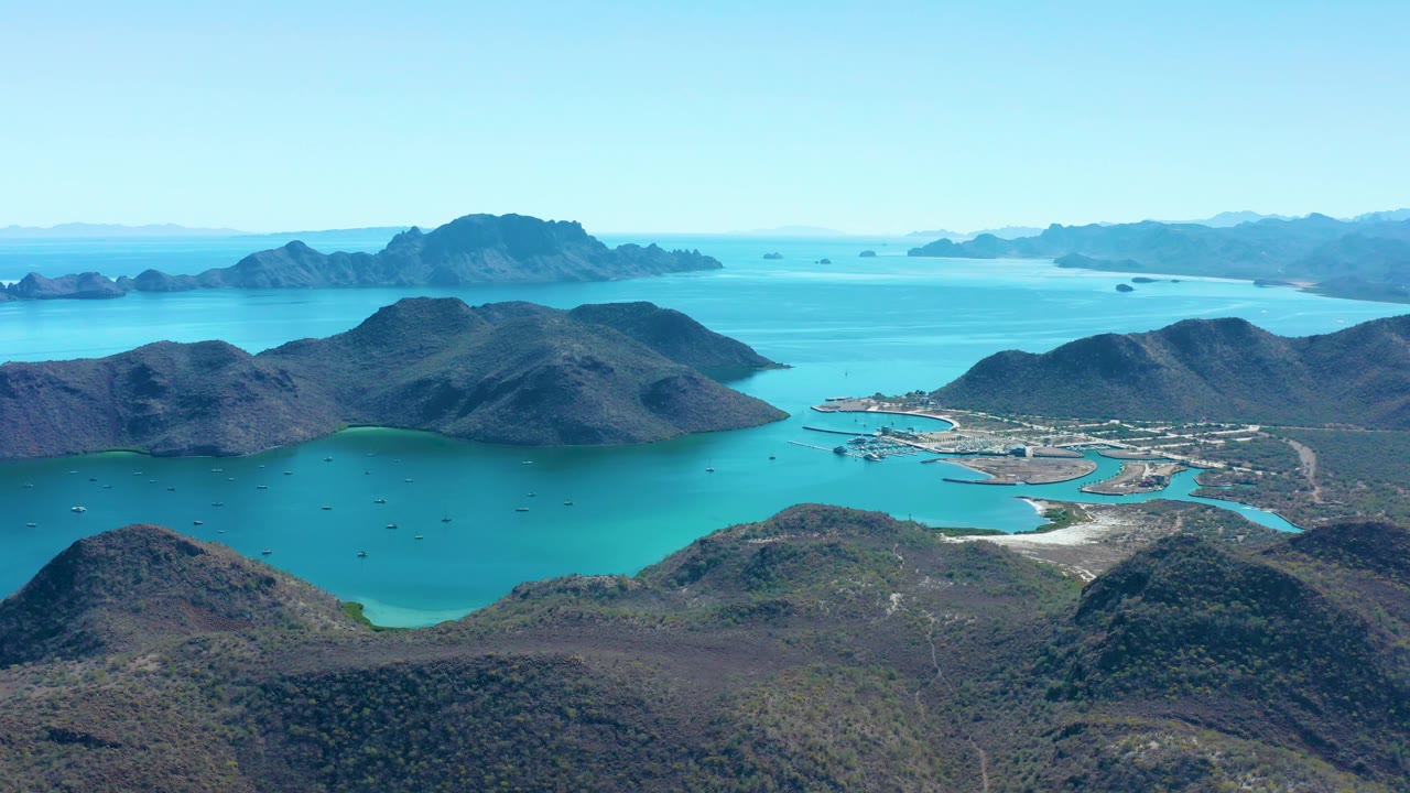 A high aerial frame of Juncalito showing the protected blue bay below rolling hills, a small developed shoreline area to the right, scattered boats on the water, and no visible people.