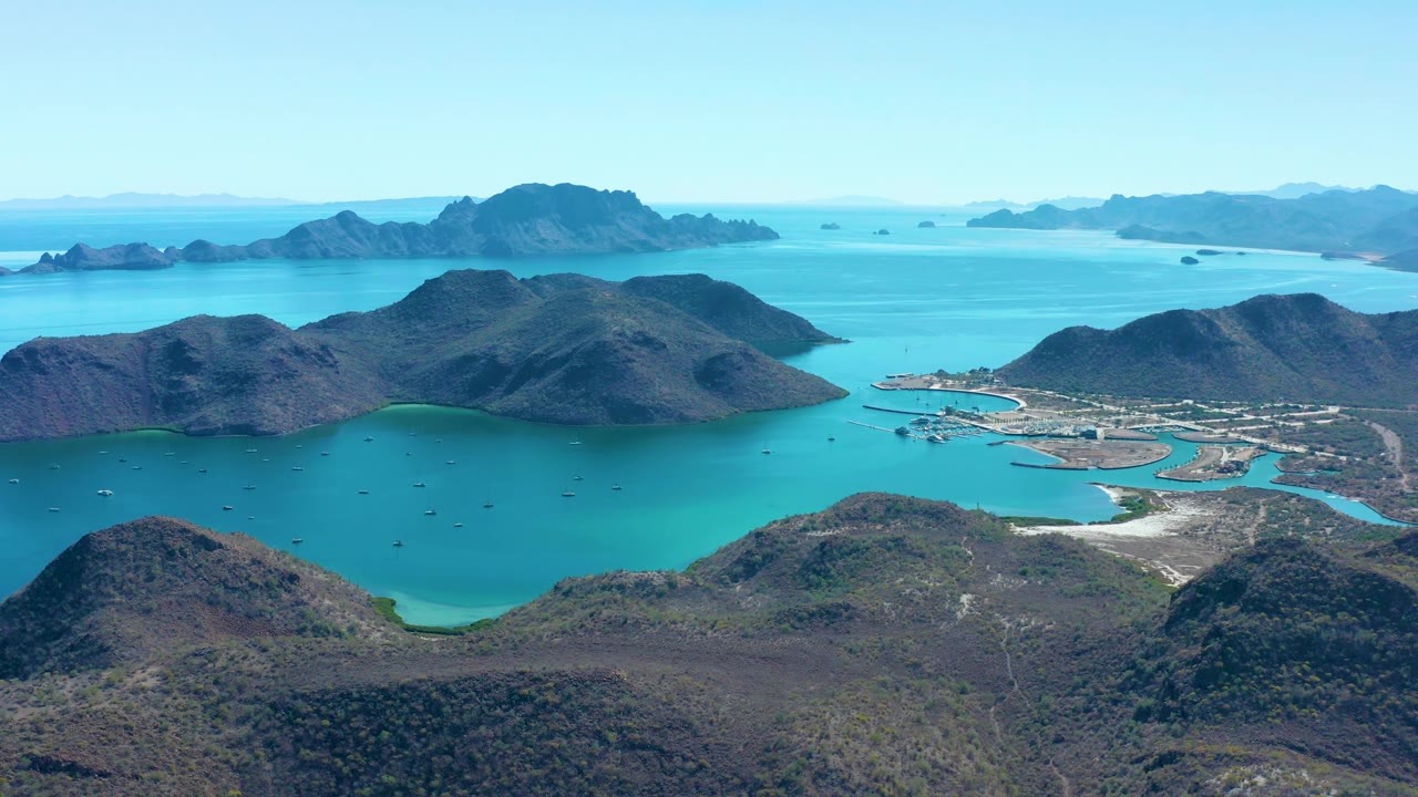 Aerial view in Juncalito, Baja California Sur, with foreground scrub-covered hills, a sheltered turquoise bay, island ridges on the horizon, small boats, and no visible people.