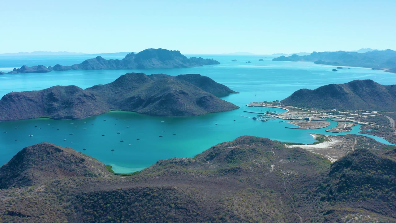 Wide drone-style frame over Juncalito, Baja California Sur, showing the bay curving around rocky hills, a small harbor area at right, distant islands, scattered boats, and no visible people.