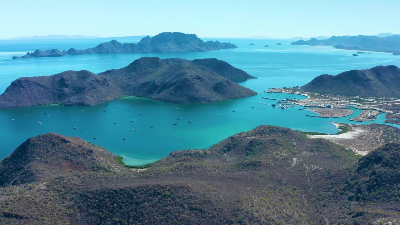 High aerial view over Juncalito, Baja California Sur, with turquoise bay water, rocky hills in the foreground, distant islands, and no visible people.