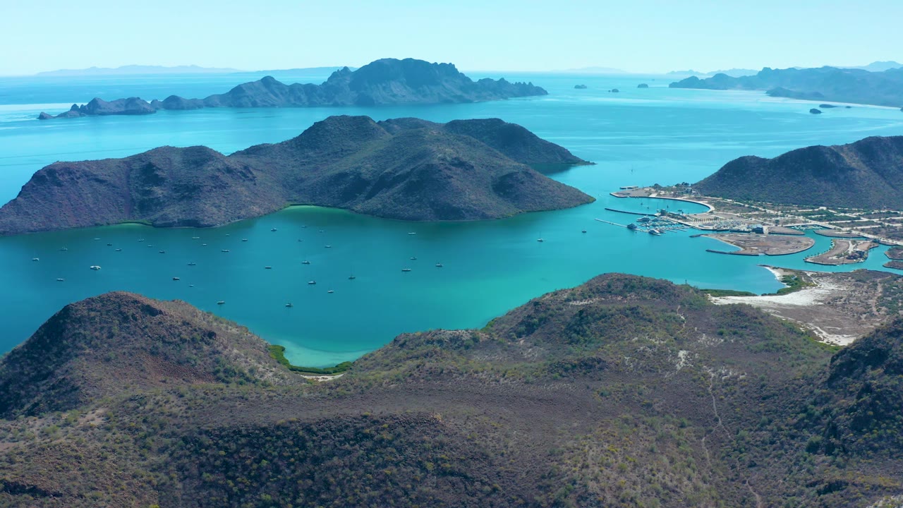 Aerial bay landscape with dry hills, turquoise water, and small boats scattered offshore.