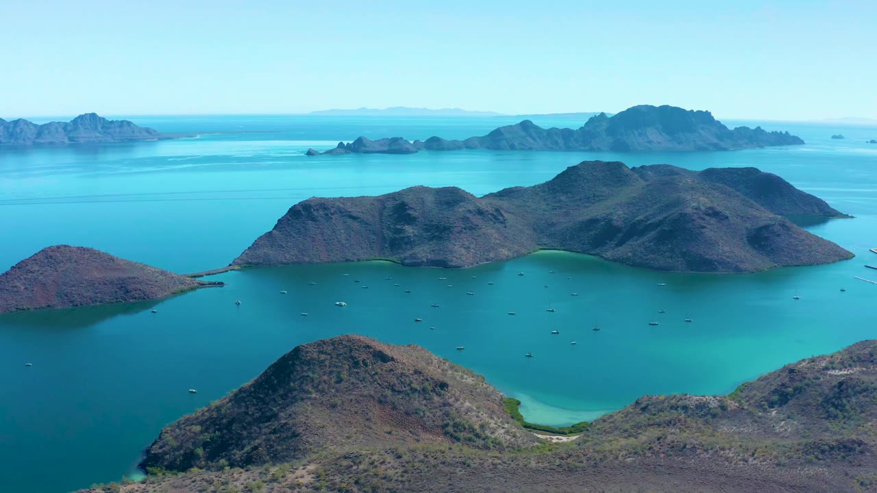Aerial frame from Juncalito with the foreground hillside lower in view, a central rocky island chain, blue-green water, small boats, and no visible people.