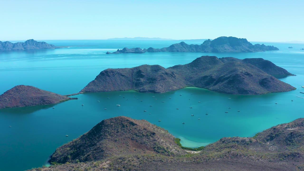 Juncalito aerial view with a bright turquoise cove below the central rocky island, darker water to the left, distant island silhouettes, and no visible people.