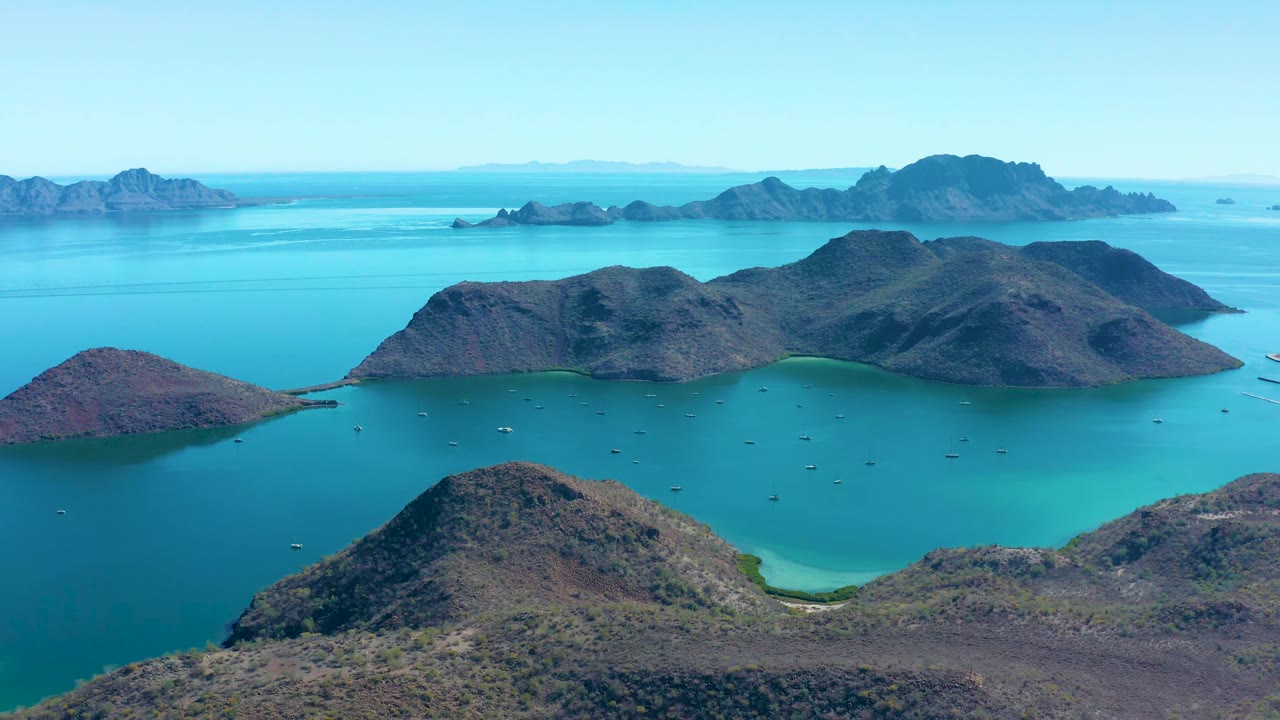 A slightly later aerial view in Juncalito shows the foreground hill below the camera, turquoise water in the cove, rocky islands across the center, and no visible people.
