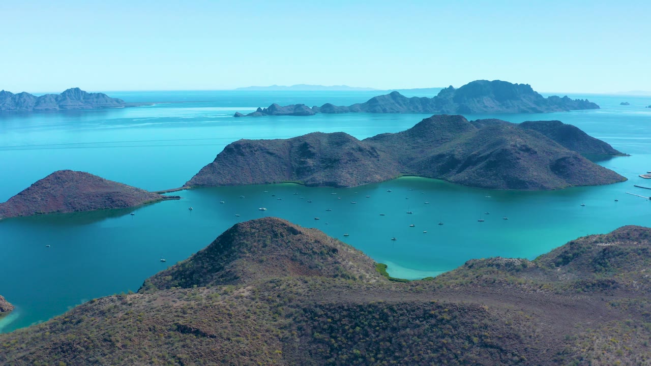 Opening aerial frame over Juncalito, Baja California Sur, showing scrub-covered rocky islands, turquoise coves, distant blue ridges, scattered small boats, and no visible people.