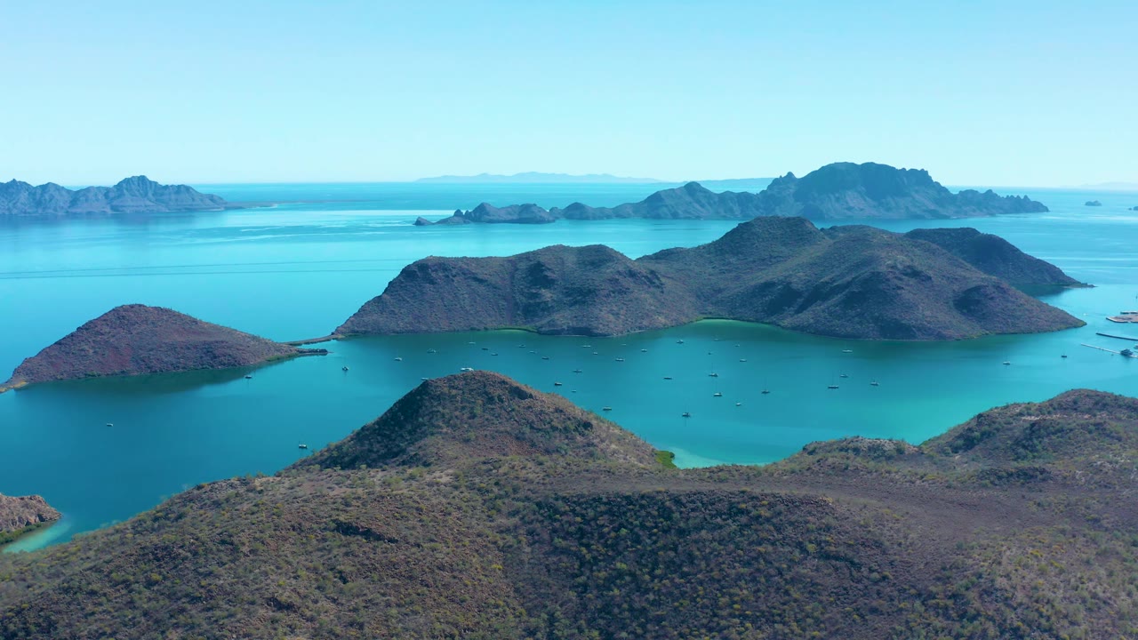 High drone frame in Juncalito, Baja California Sur, looking across a foreground hill toward blue-green water, low rocky islands, distant ridges, and no visible people.