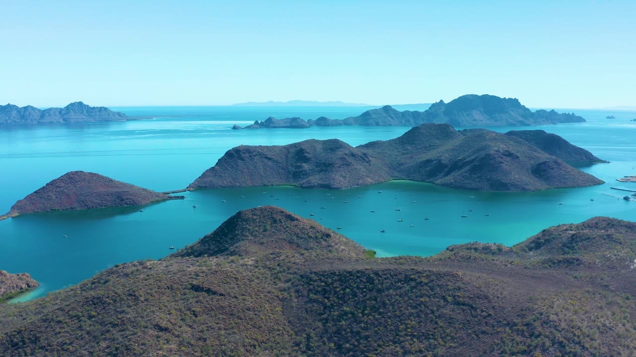 Mid-clip aerial view in Juncalito, Baja California Sur, showing rugged brown hills around a turquoise bay, layered island silhouettes, open sky, and no visible people.