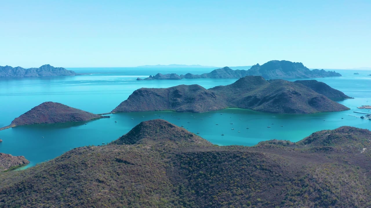 Aerial view over low dark island hills near Juncalito, Baja California Sur, Mexico, with blue-green channels, faint boats in the bay, and no visible people.