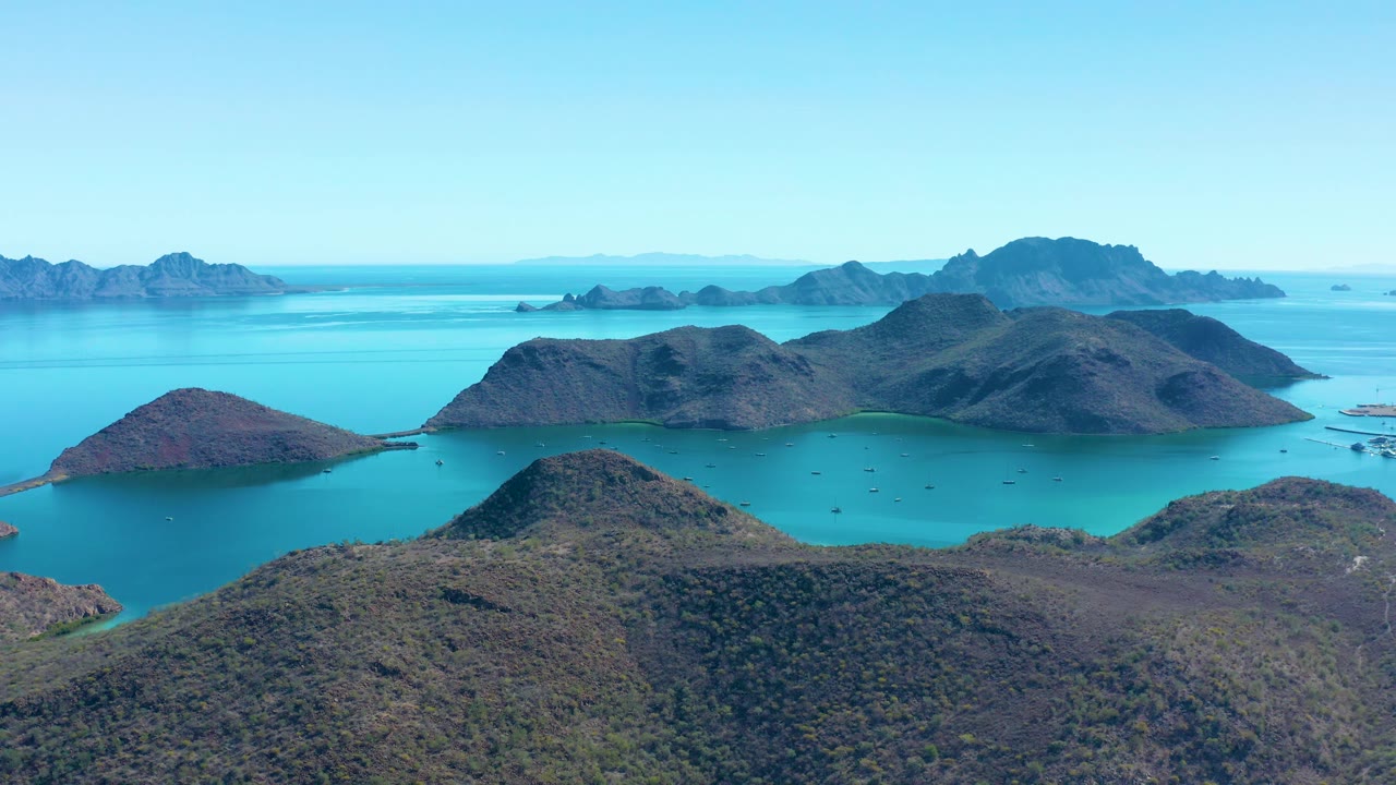 Wide coastal aerial near Juncalito, Baja California Sur, Mexico, showing island hills, turquoise bay water, faint boats to the right, and no visible people.
