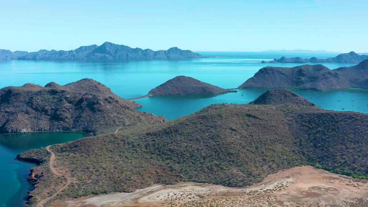 Aerial frame from Juncalito, Baja California Sur showing scrub-covered hills, turquoise bays, distant mountain ranges, pale shoreline terrain, and no visible people.