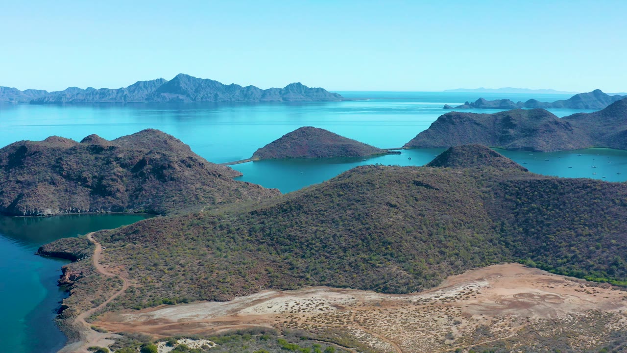 A wide aerial frame in Juncalito shows the winding shore road lower left, scrub-covered hills across the center, turquoise bays behind them, mountains on the horizon, and no visible people.