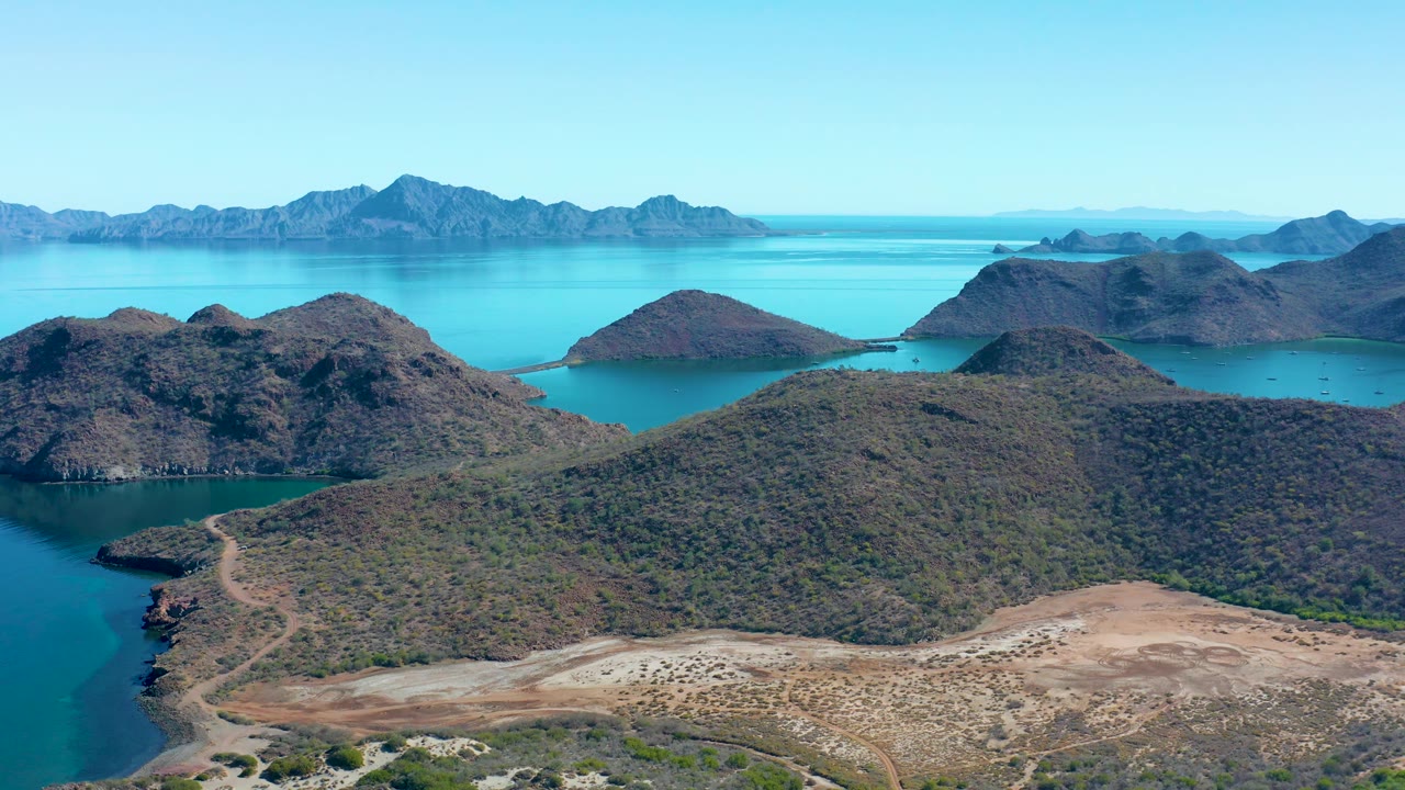 At 0:03, the frame shows more of the dry shoreline foreground beneath Juncalito's coastal hills, with the winding road, bright turquoise water, hazy distant mountains, and no visible people.
