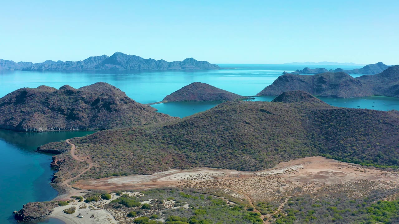 At 0:01, the Juncalito coastline remains in a high wide view, with rocky hills above a pale sandy waterline, turquoise inlet water, distant island ridges, and no visible people.