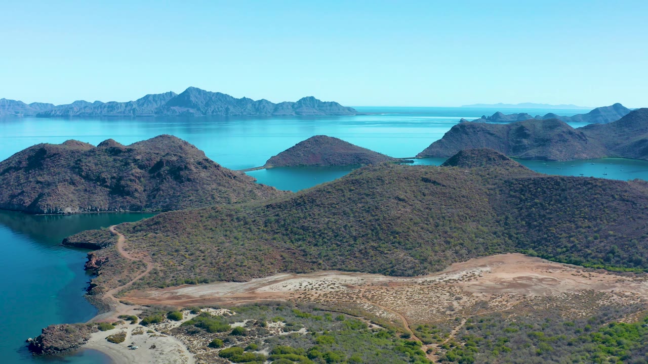 At 0:02, scrub-covered hills dominate the Juncalito foreground while a sandy coastal edge meets calm turquoise water, with layered island mountains on the horizon and no visible people.